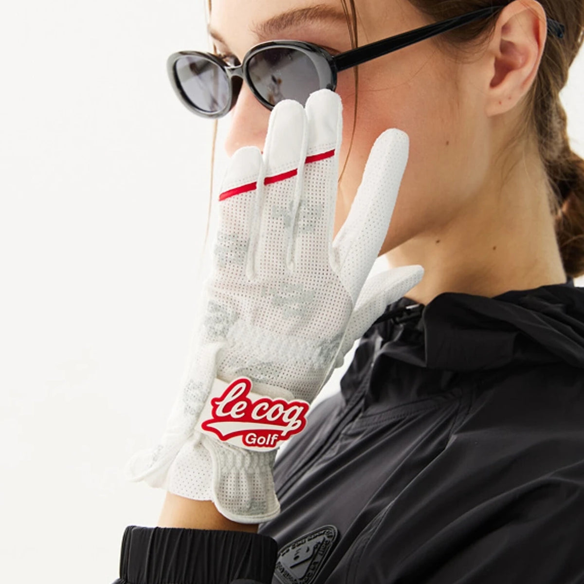 A close-up lifestyle shot of a model wearing a white Le Coq Golf glove with red accents and sunglasses, paired with a black jacket against a clean white background.