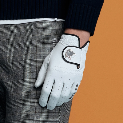 A white leather golf glove with black trim and a gray bird logo, worn on a hand over a gray plaid trouser leg, against an orange background.