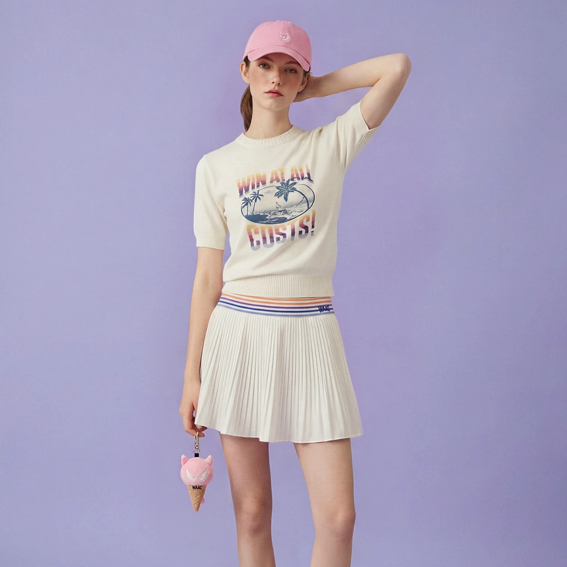 A model wearing a cream-colored short-sleeve knit top with a colorful graphic print, a matching pleated mini skirt with a striped waistband, and a pink baseball cap, standing against a solid lavender background.