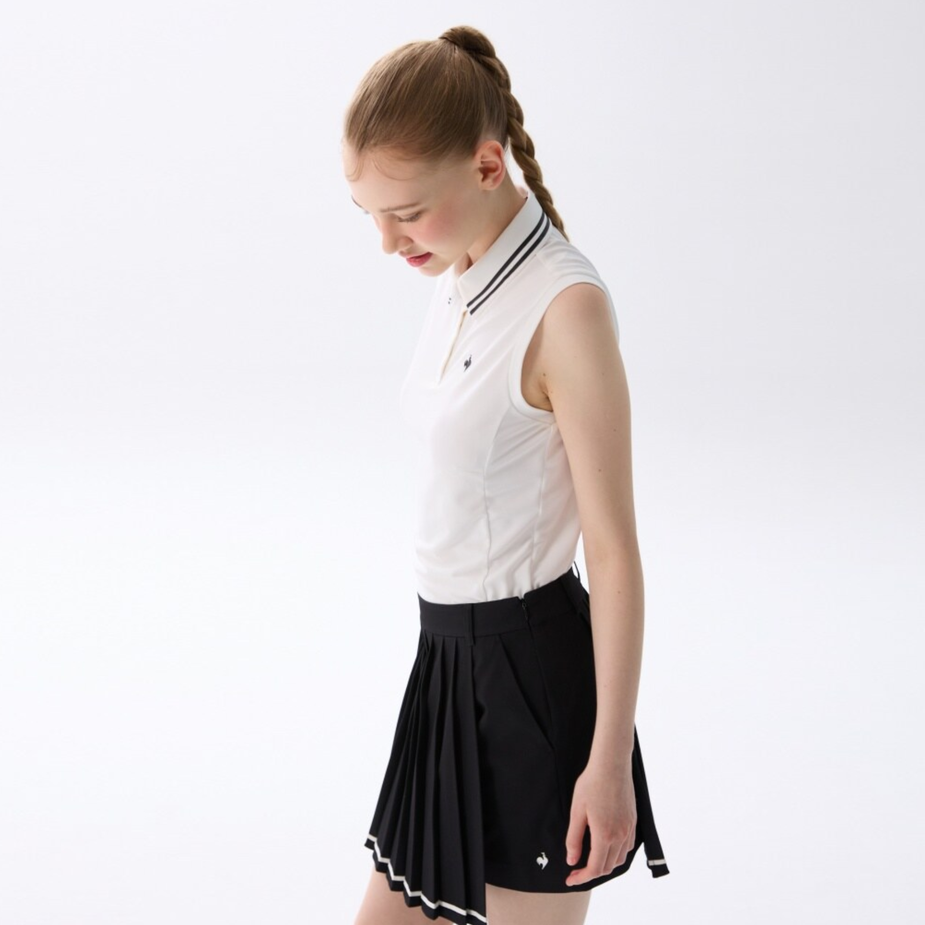 A young woman with her hair in a single braid is shown from the side, wearing a white sleeveless polo shirt with a black and white striped collar, paired with a black pleated tennis skirt featuring white trim.