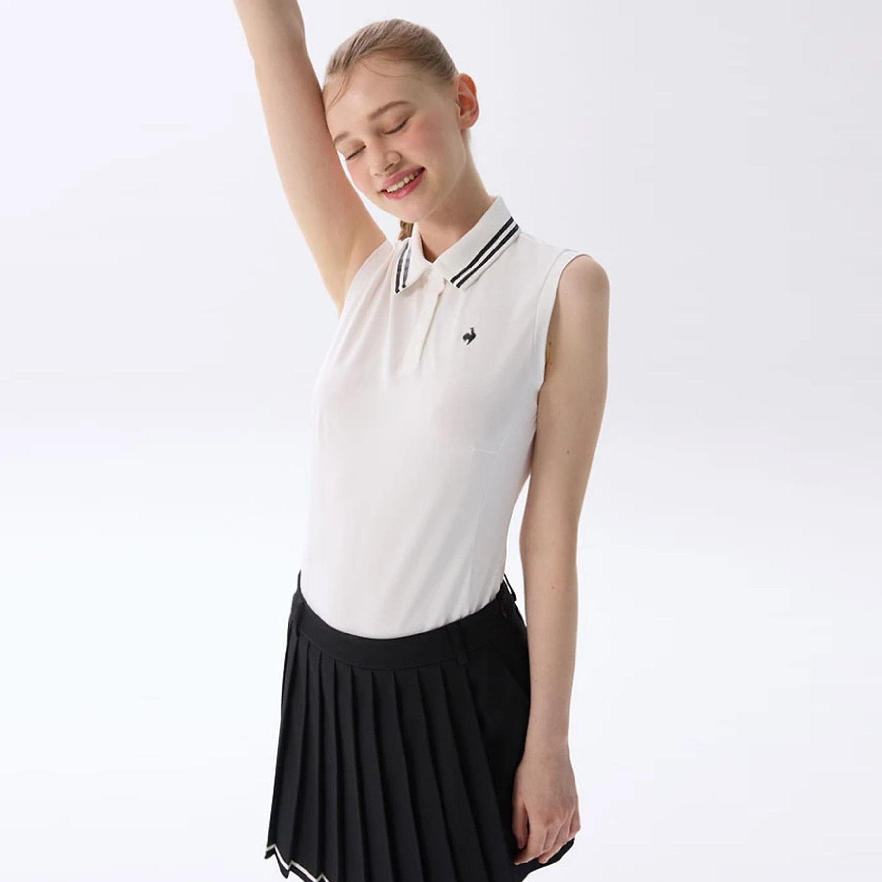 A young girl modeling a sleeveless white polo shirt with black trim on the collar and a black pleated skirt, posed against a clean white studio background.
