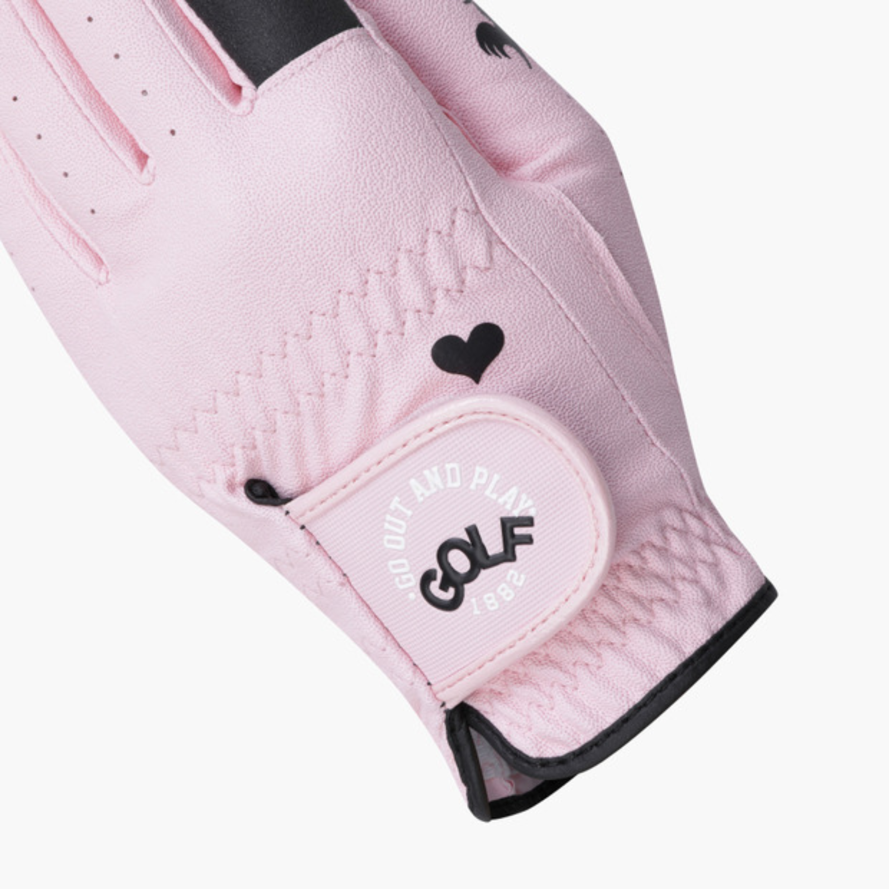 A close-up of a light pink golf glove with black accents, including a heart detail and the 'GO OUT AND PLAY GOLF' logo on the wrist strap, set against a plain light gray background.
