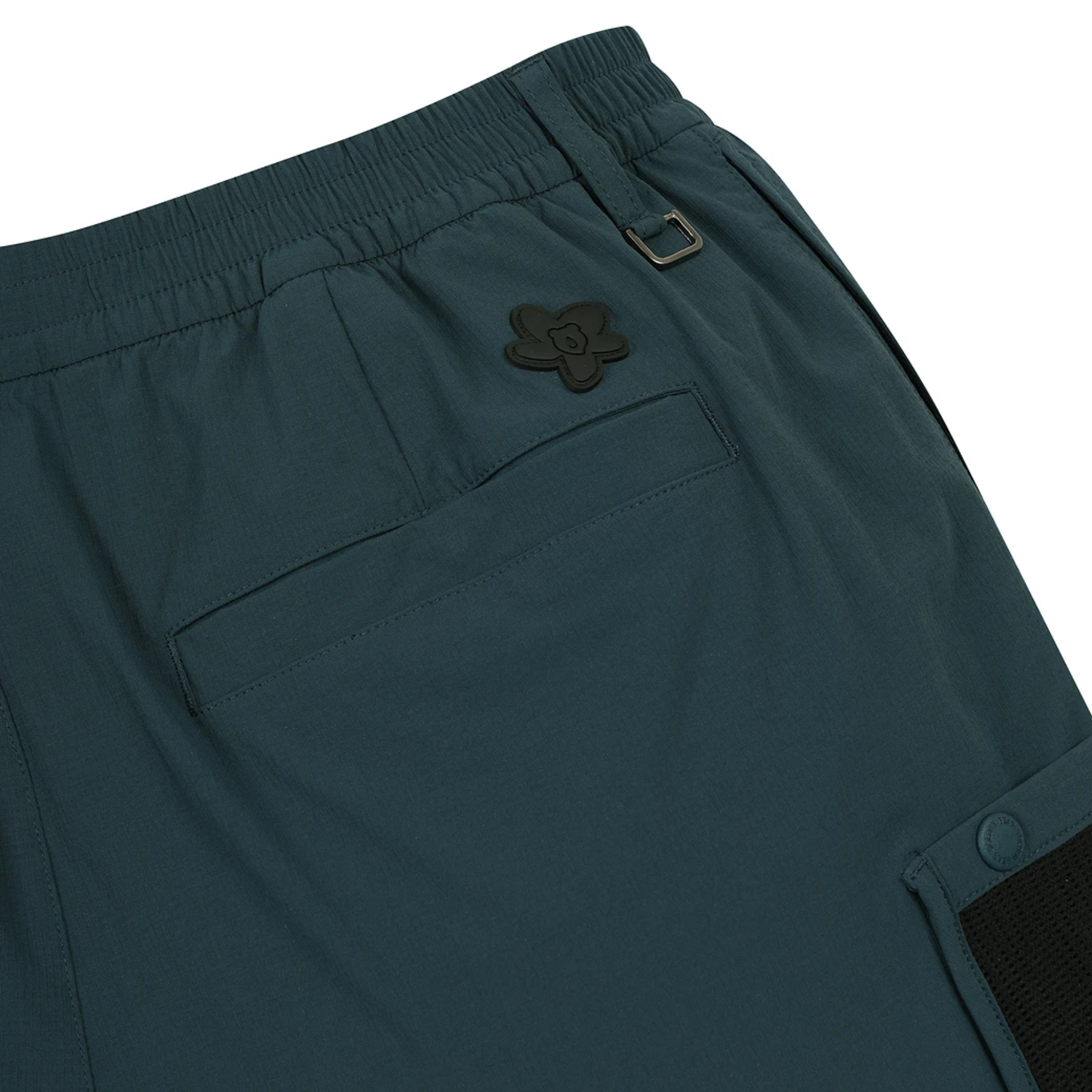 Close-up of dark teal outdoor shorts showing the elastic waistband, a side pocket with a dark logo patch, and a cargo pocket with a snap closure on a white background.