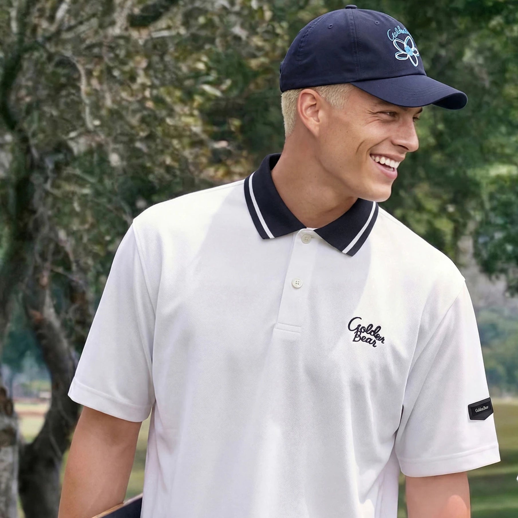 A man wearing a white polo shirt with a navy blue collar and embroidered 'Golden Bear' logo on the chest, paired with a navy blue baseball cap, set against a blurred outdoor background of trees.