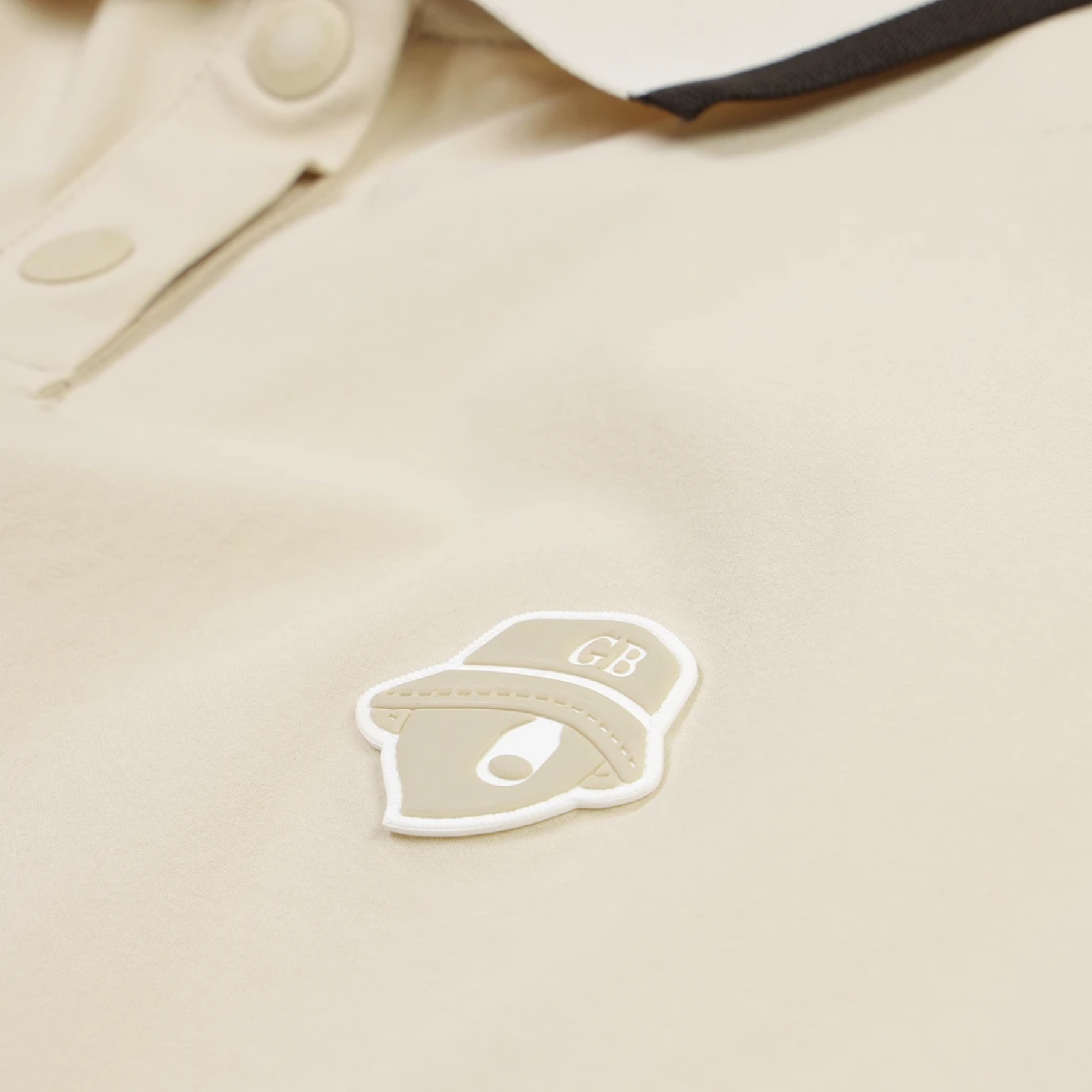 Close-up of a beige polo shirt with a small shield-shaped patch featuring the letters GB and a stylized eye logo, plus part of a dark collar stripe.