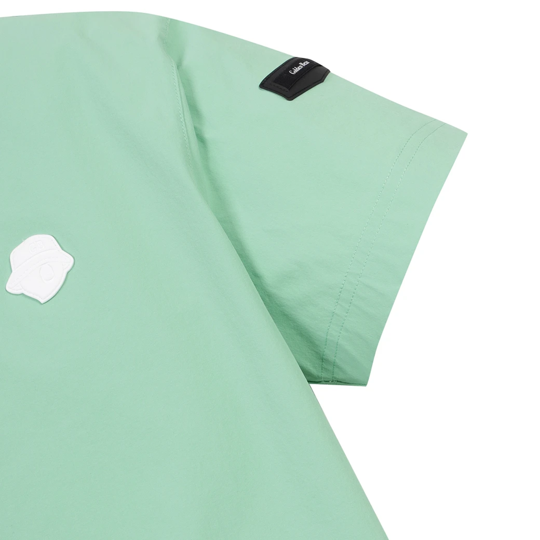 A close-up of a mint green scrub top showing the sleeve, shoulder seam, a small black brand patch, and a white shield-shaped patch on the chest, set against a plain white background.
