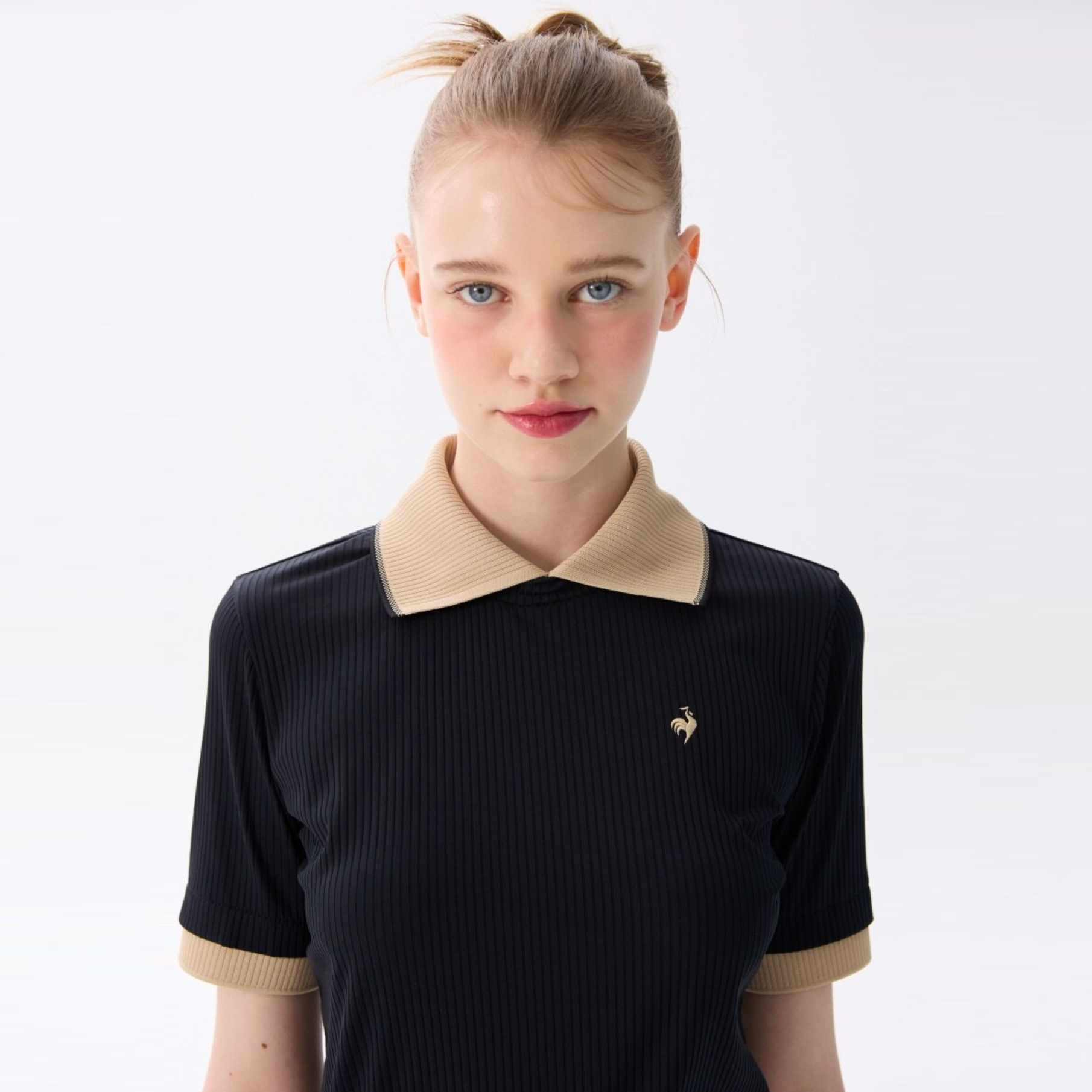 A young woman with blonde hair in a high ponytail wearing a black ribbed short-sleeve polo shirt with a beige collar and cuffs, featuring a small gold rooster logo on the chest, set against a plain white studio background.