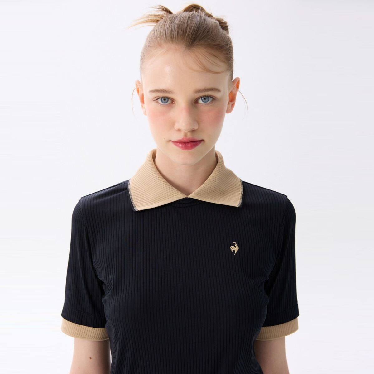 A young woman with blonde hair in a high ponytail wearing a black ribbed short-sleeve polo shirt with a beige collar and cuffs, featuring a small gold rooster logo on the chest, set against a plain white studio background.