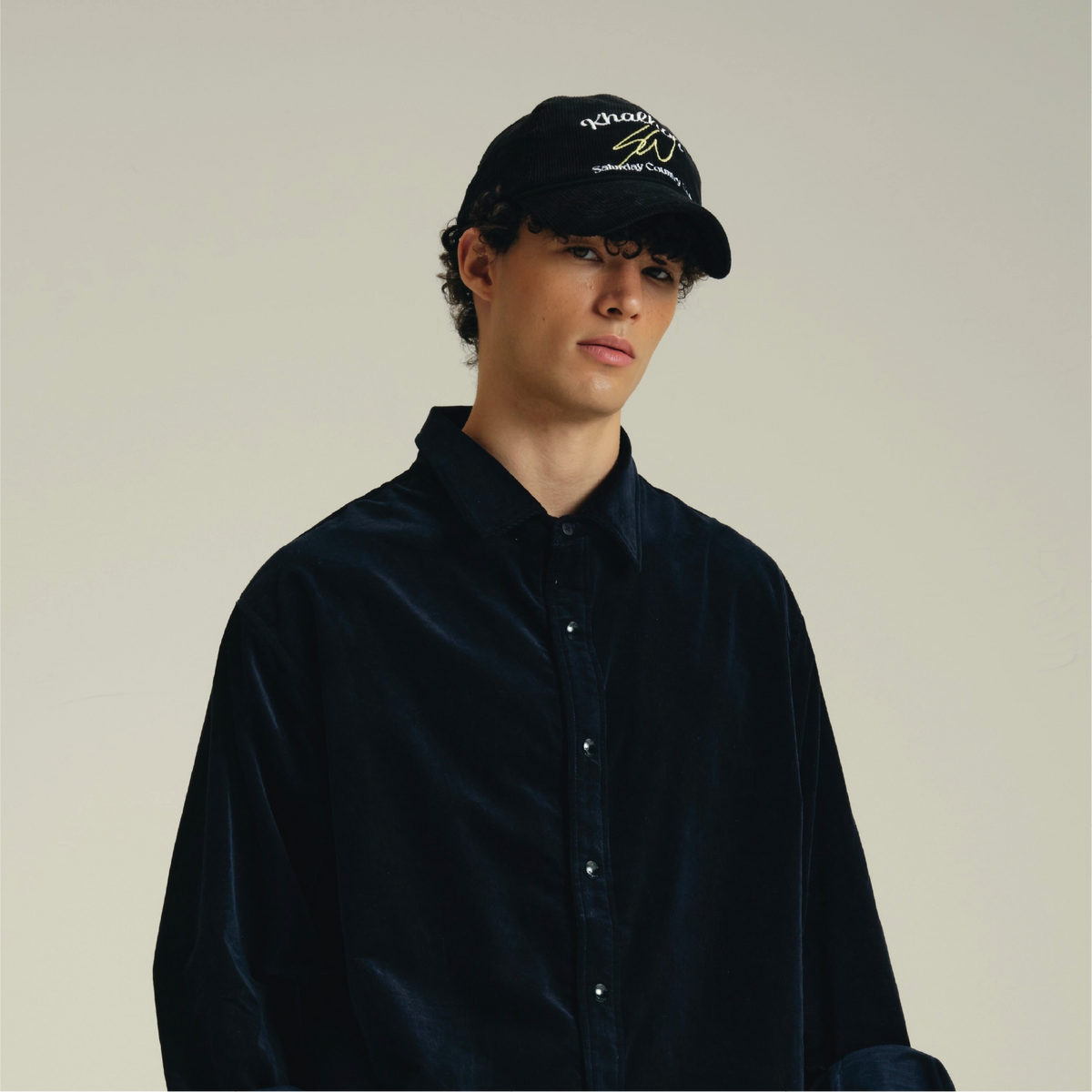 A model wearing a black velvet button-up shirt and a black baseball cap with yellow and white embroidered text, photographed against a solid light beige background.