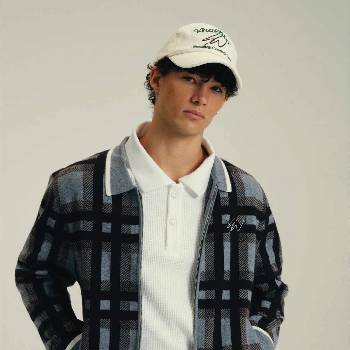 A model wearing a white baseball cap with green embroidery, a white ribbed polo shirt, and a gray and black plaid zip-up jacket, photographed against a solid light gray background.