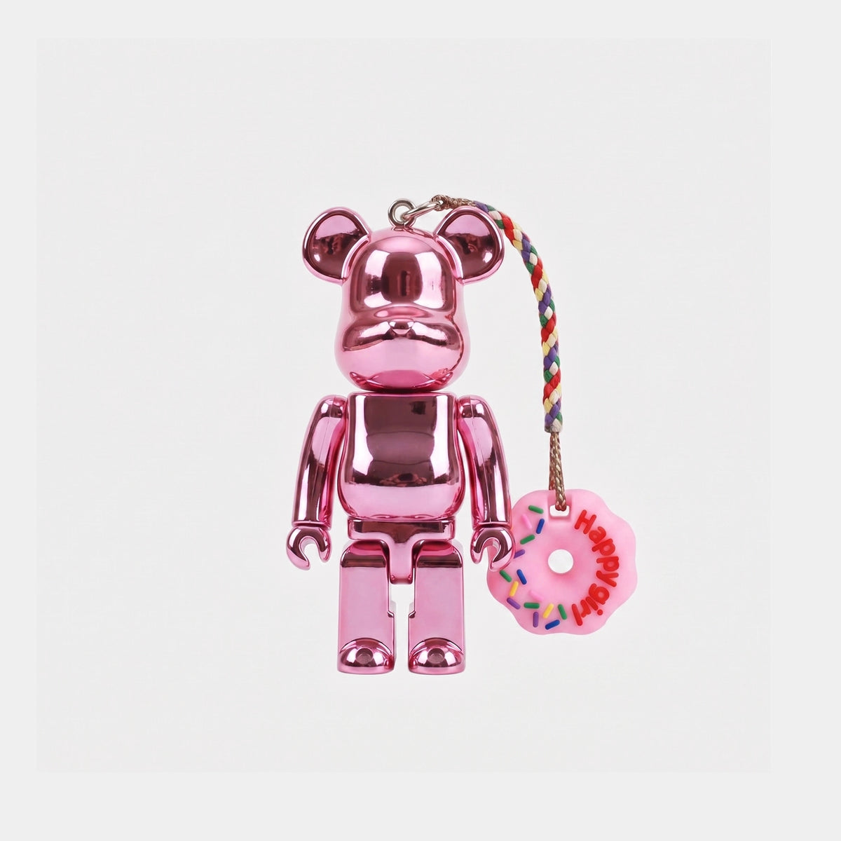 A metallic pink Bearbrick keychain featuring a shiny pink bear figure with a pink donut charm hanging from a multicolor braided cord. The donut charm is decorated with colorful sprinkles and the word 'Happy' in red.