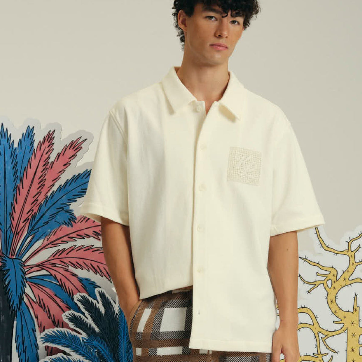 A man wearing a short-sleeve cream-colored button-up shirt with a textured square patch on the left chest, paired with brown and white plaid trousers, standing against a light beige wall with colorful tropical cutout decorations.