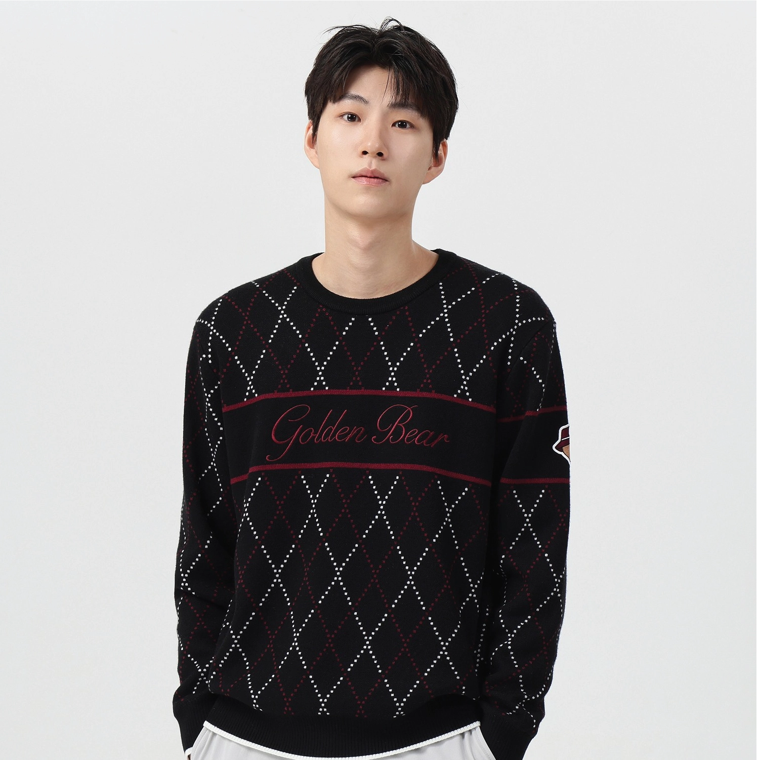 A man wearing a black crewneck sweater with a white and red argyle pattern and 'Golden Bear' text across the chest, paired with white pants, against a light gray background.
