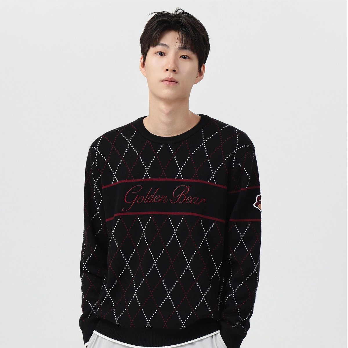A man wearing a black crewneck sweater with a white and red argyle pattern and 'Golden Bear' text across the chest, paired with white pants, against a light gray background.