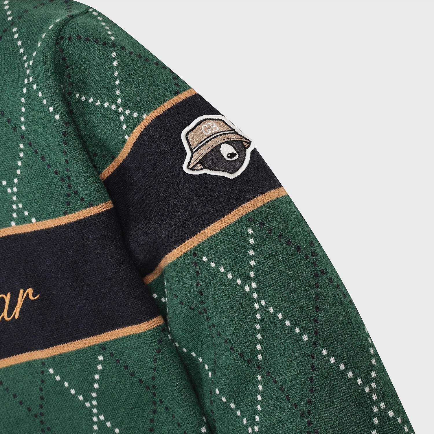 Close-up of a green patterned knit garment with dark navy bands, tan trim, and a small embroidered raccoon hat patch on the sleeve.