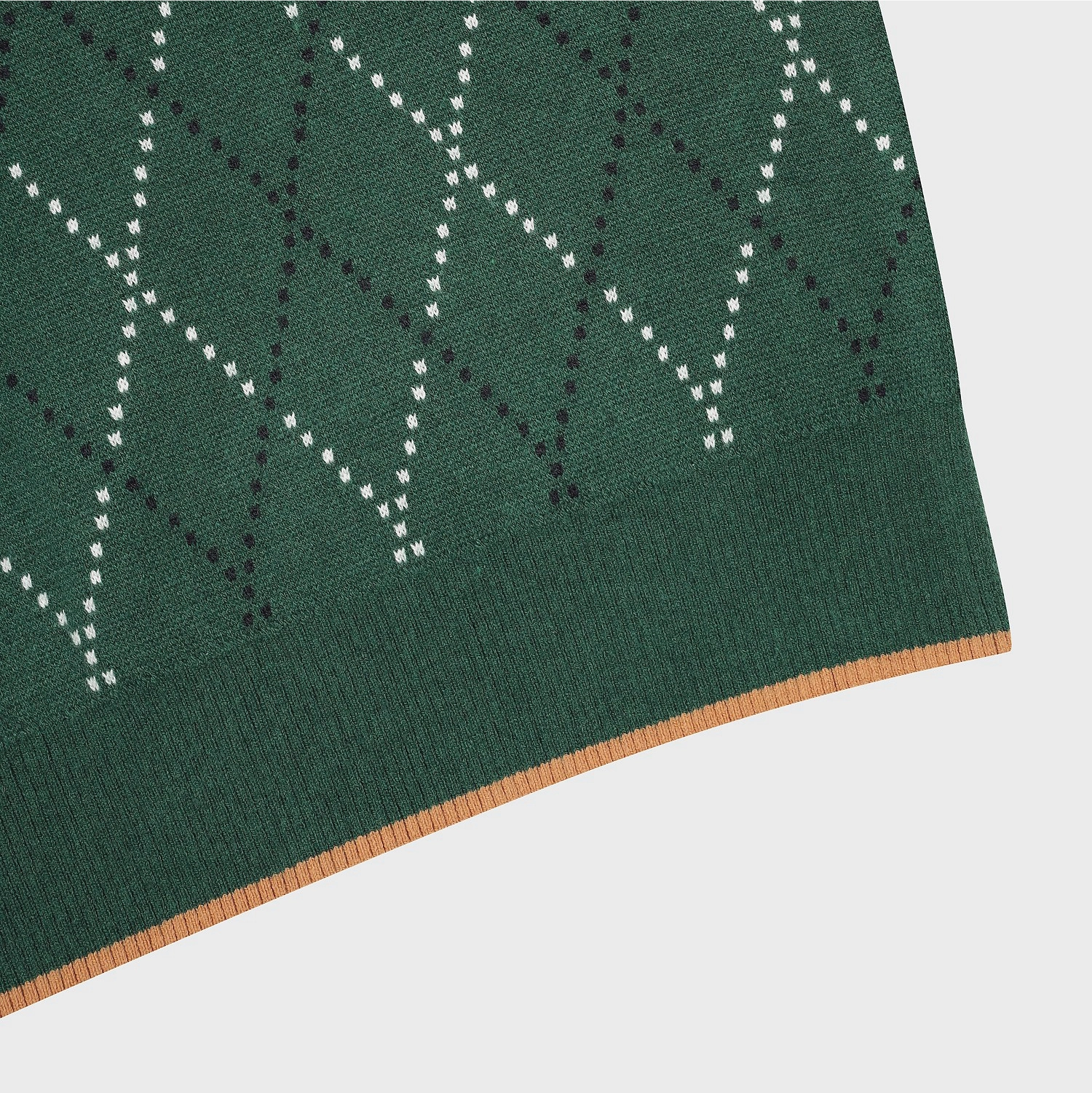 Close-up of a knitted textile corner showing a dark green base with a white and black dotted geometric pattern and a tan stitched edge on a light gray background.