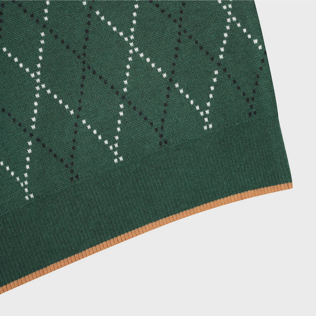 Close-up of a knitted textile corner showing a dark green base with a white and black dotted geometric pattern and a tan stitched edge on a light gray background.