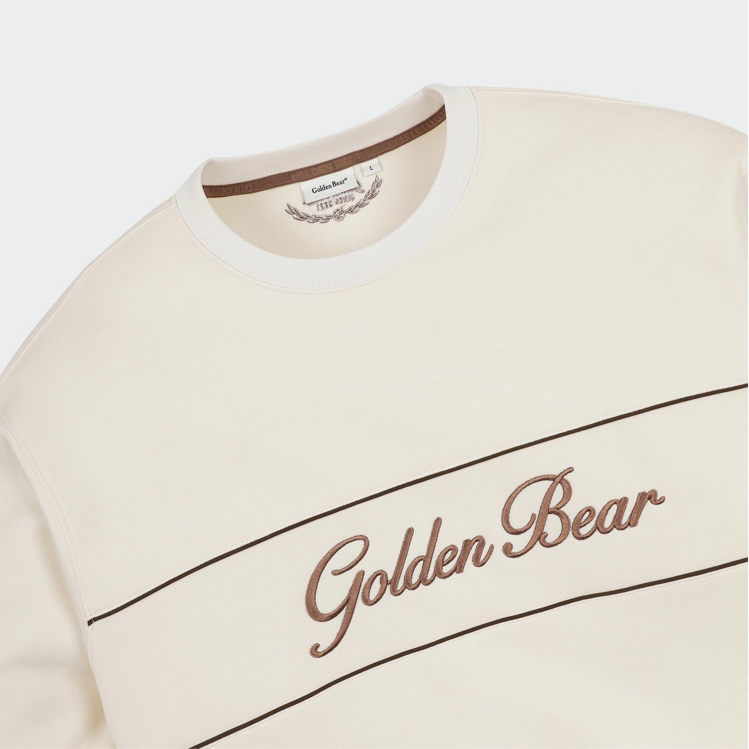 Close-up of a cream-colored crewneck sweatshirt featuring brown embroidered 'Golden Bear' script across a horizontal stripe, with a matching inner collar label.