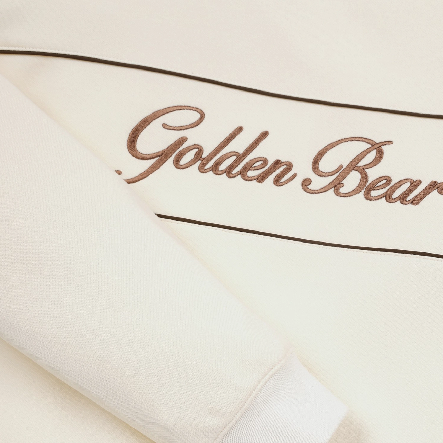 Close-up of a cream-colored sweatshirt featuring metallic bronze cursive embroidery that reads 'Golden Bear' between two thin dark brown horizontal lines.