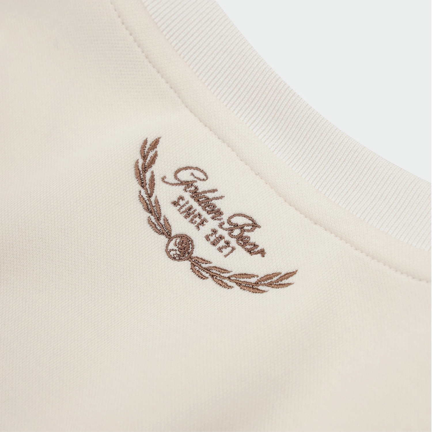 Close-up of a cream-colored ribbed knit garment with a brown embroidered logo reading 'Golden Bear' and 'SINCE 2021' on the inside neckline.