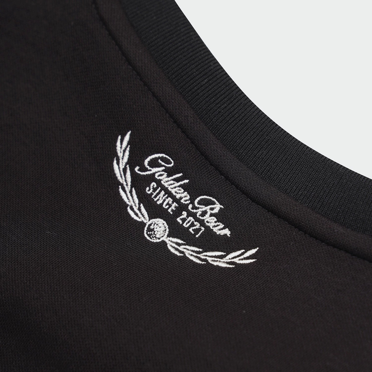 Close-up of a black crewneck t-shirt showing a white embroidered logo on the inside collar that reads 'Golden Bear SINGE 2021' with a laurel wreath.