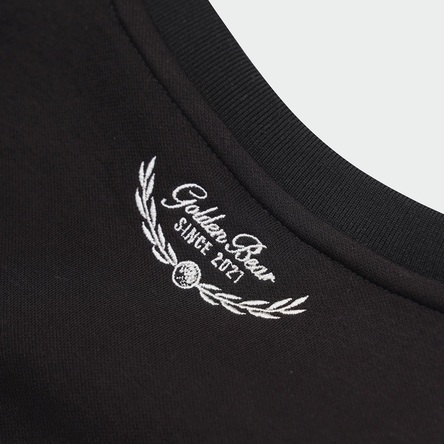 Close-up of a black crewneck t-shirt showing a white embroidered logo on the inside collar that reads 'Golden Bear SINGE 2021' with a laurel wreath.