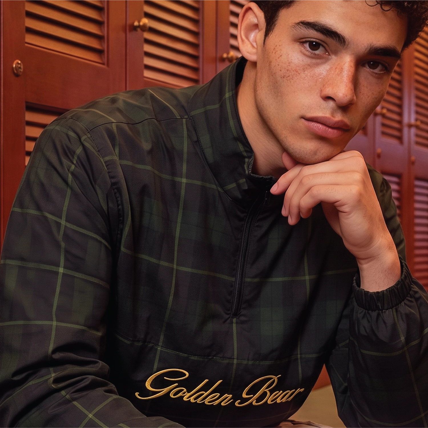 A man wearing a dark green and black plaid quarter-zip pullover jacket with 'Golden Bear' embroidered in gold script on the chest, posed in front of wooden louvered doors.