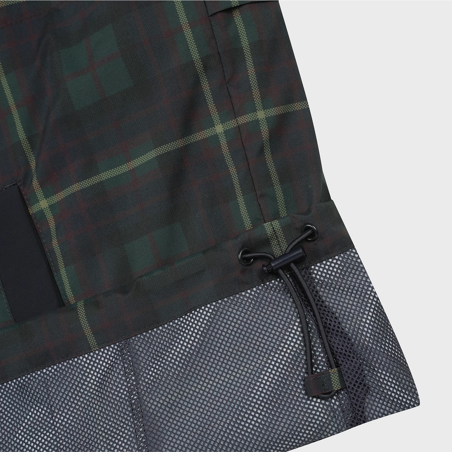 Close-up of a plaid garment, likely a jacket or coat, showing a dark green and black tartan pattern with thin yellow-green lines, a black drawcord, and a dark gray mesh hem panel on a light gray background.