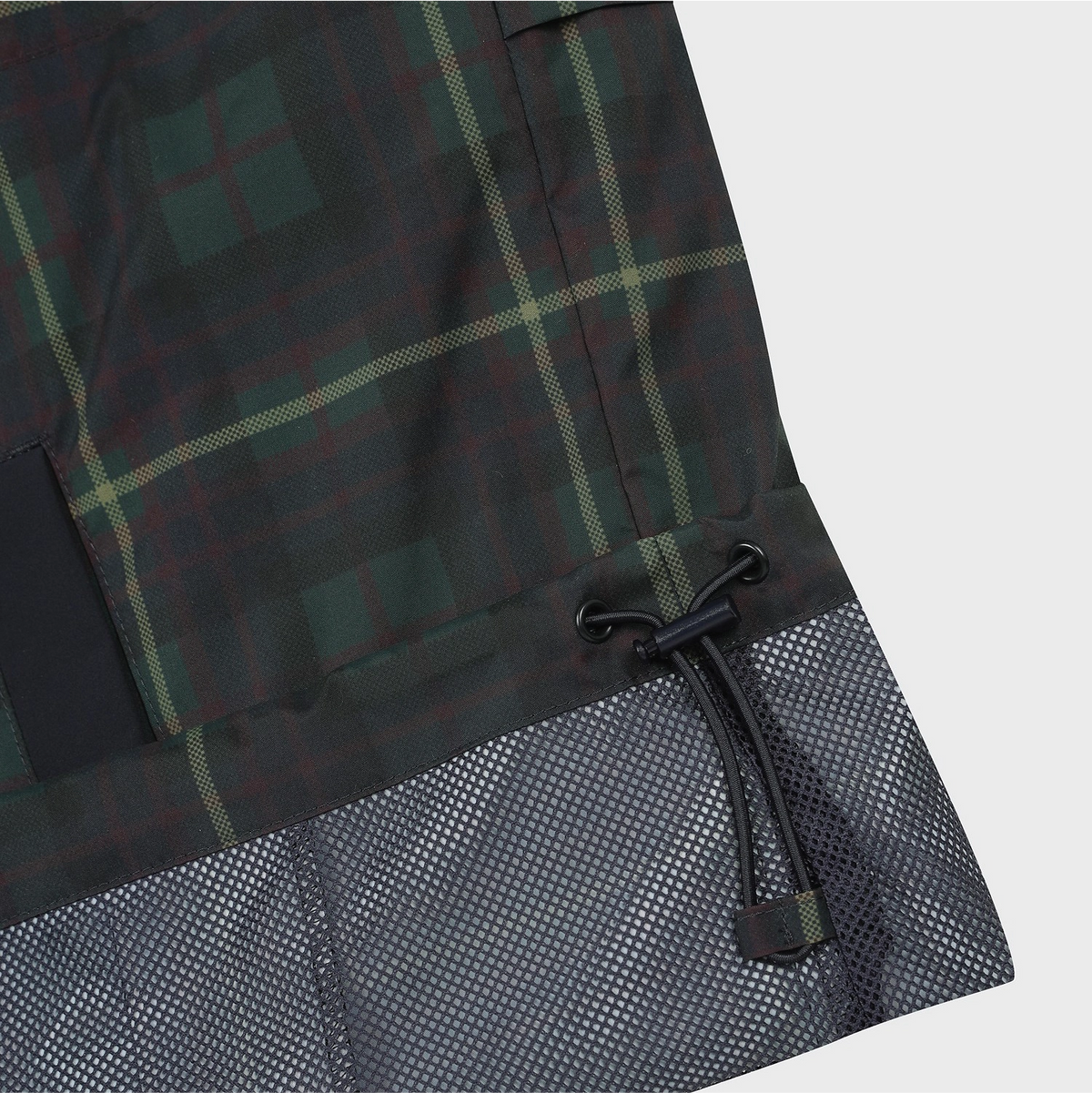 Close-up of a plaid garment, likely a jacket or coat, showing a dark green and black tartan pattern with thin yellow-green lines, a black drawcord, and a dark gray mesh hem panel on a light gray background.