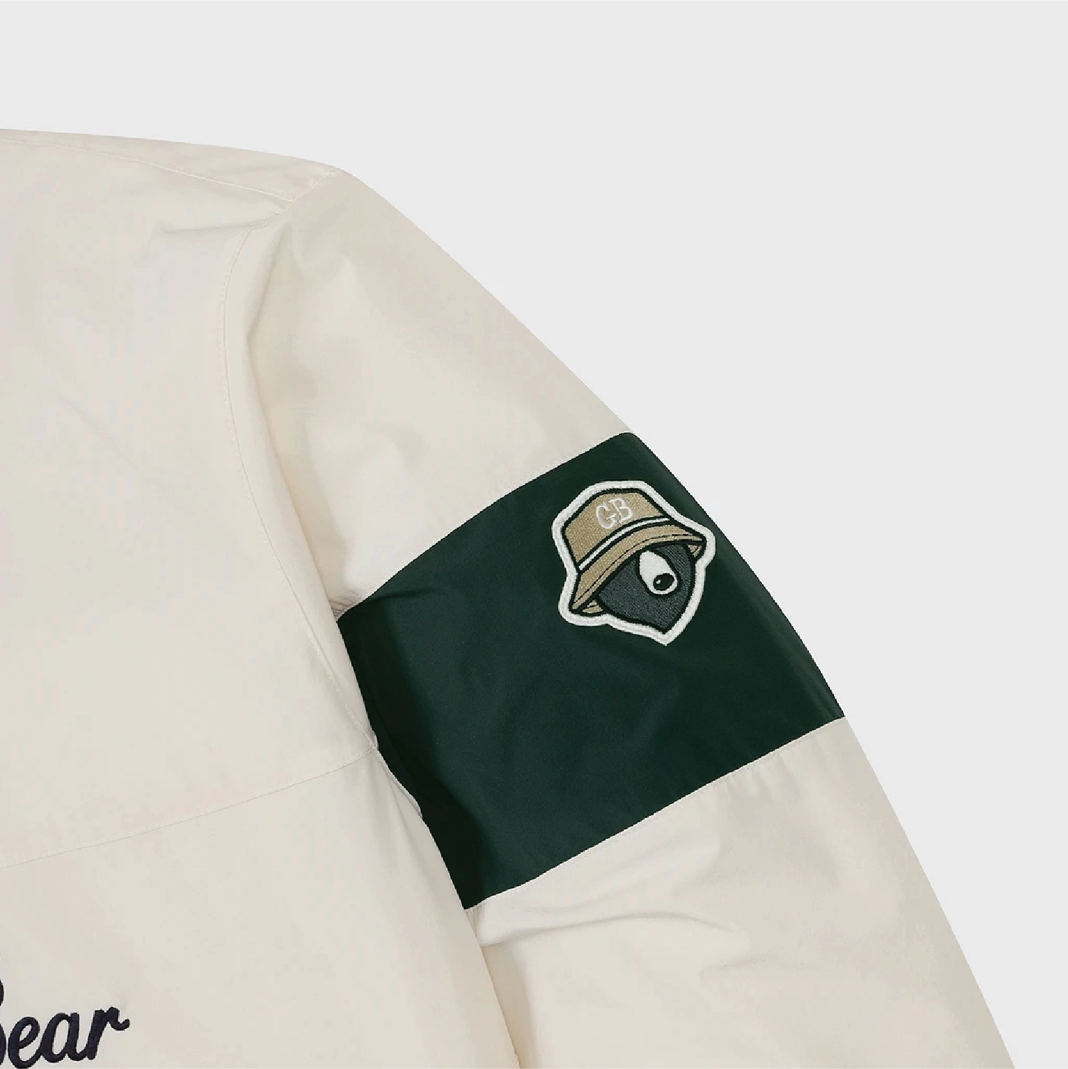 Close-up of a jacket sleeve with an off-white body, a dark green banded cuff, and a patch featuring a stylized bear face wearing a hat.