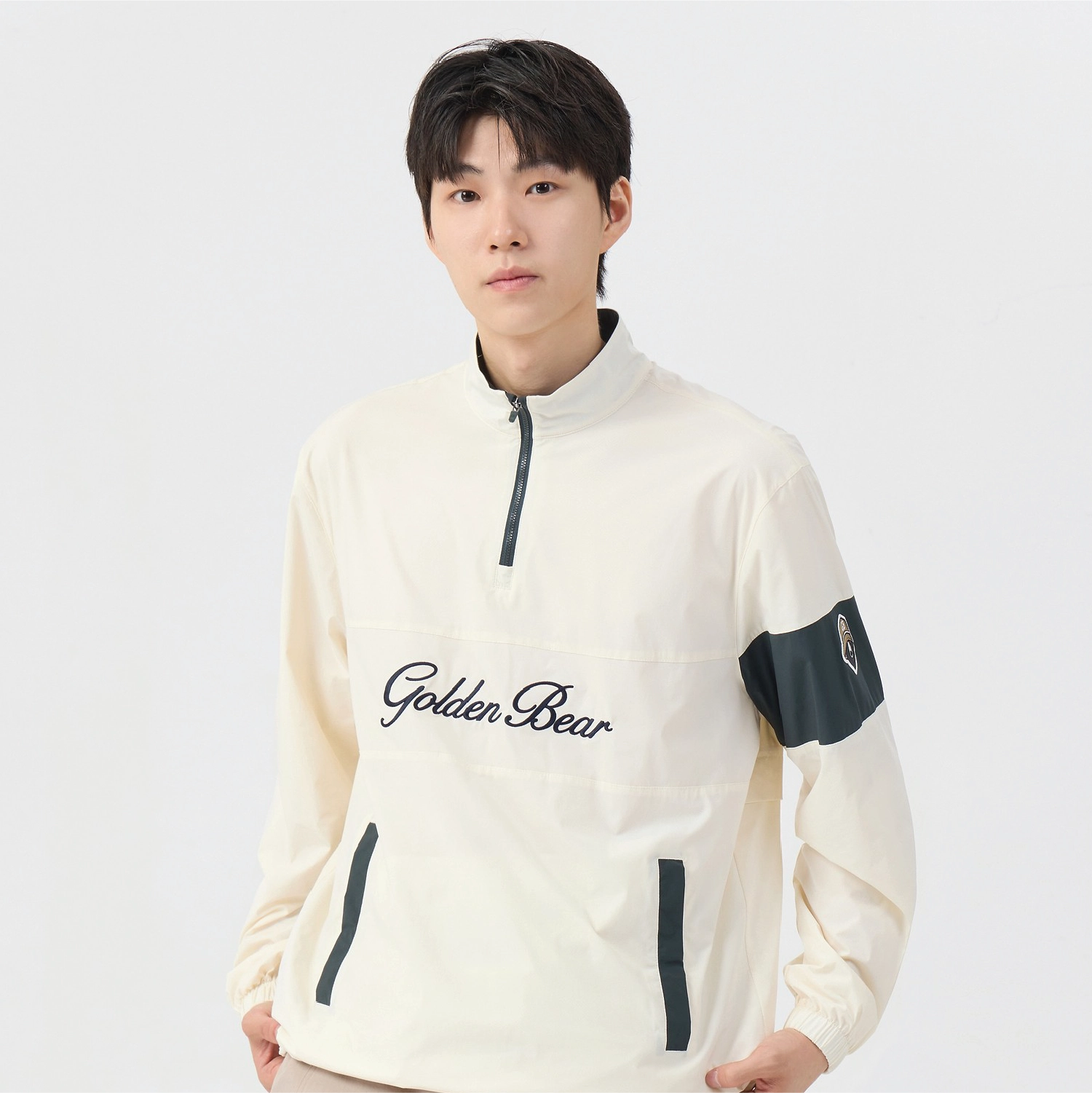 A young man wearing a cream-colored quarter-zip pullover jacket with 'Golden Bear' embroidered in black script across the chest, dark green contrast piping, and a dark green sleeve patch.