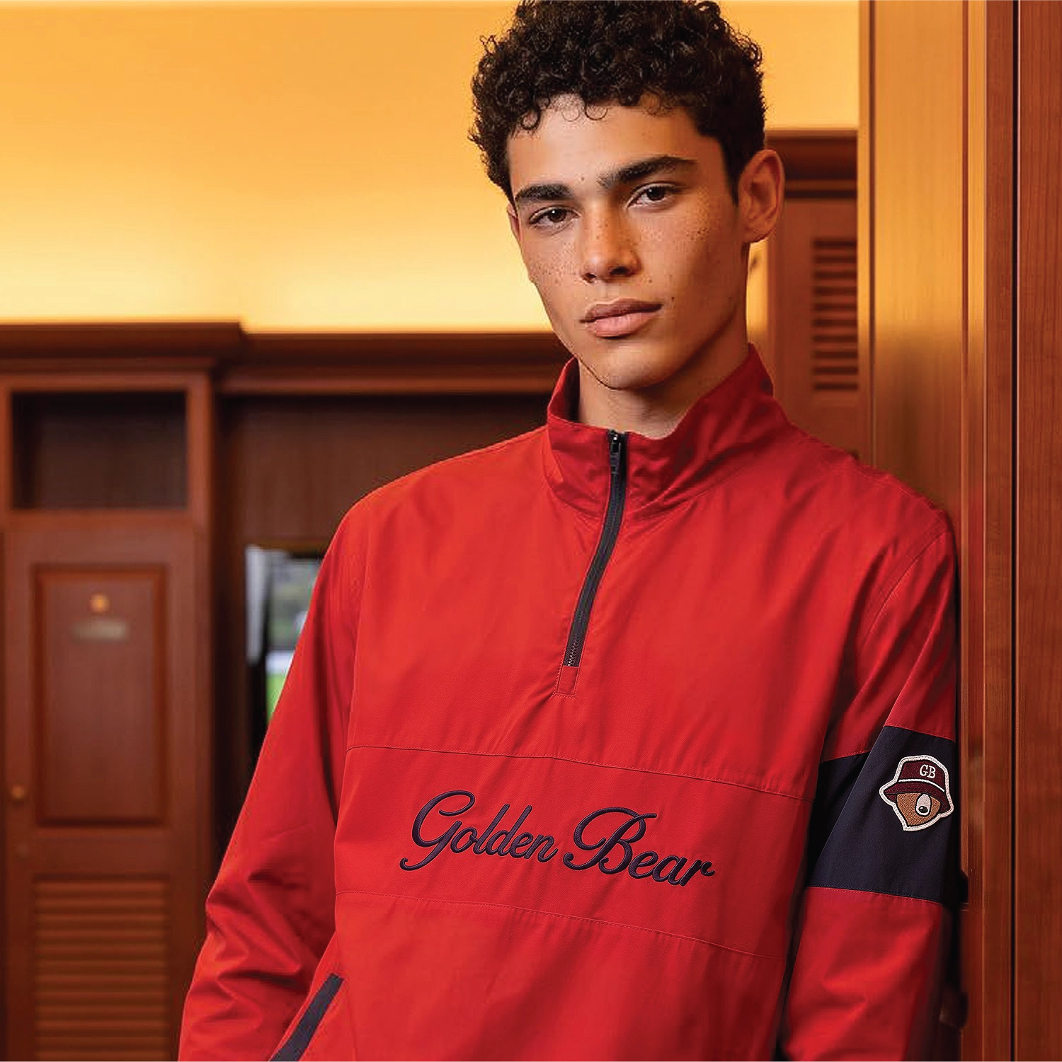 A young man with curly dark hair stands in a locker room wearing a bright red quarter-zip pullover with 'Golden Bear' embroidered across the chest and a dark navy sleeve patch.