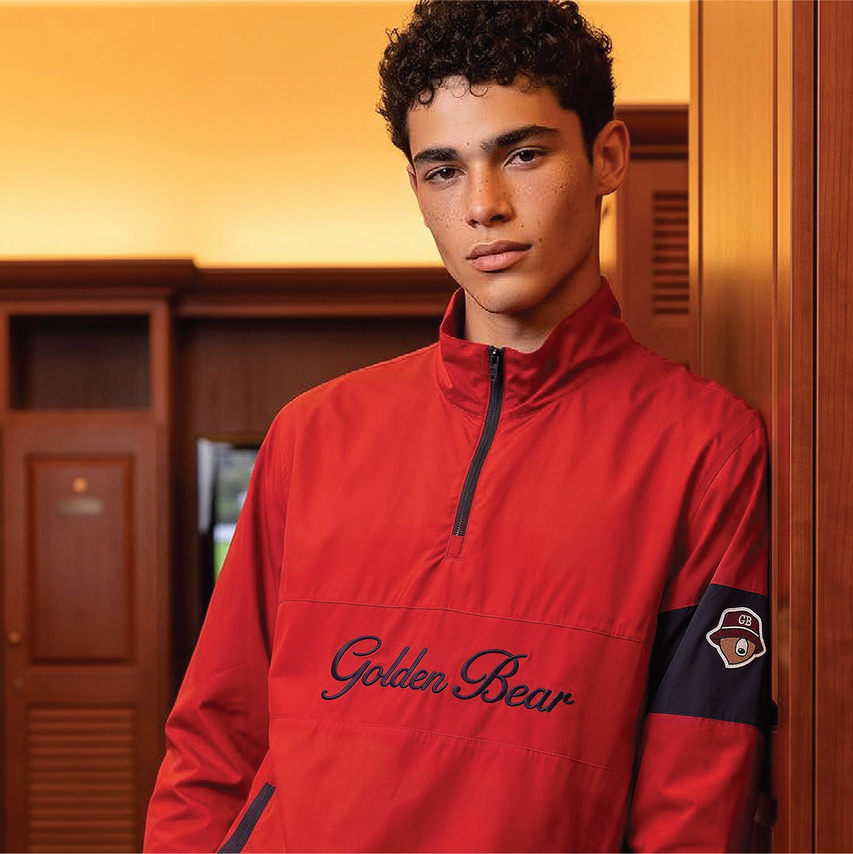 A young man with curly dark hair stands in a locker room wearing a bright red quarter-zip pullover with 'Golden Bear' embroidered across the chest and a dark navy sleeve patch.