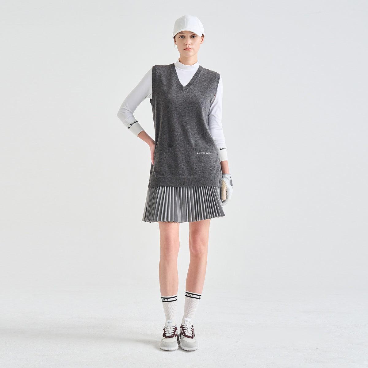 A model stands against a light gray studio background wearing a gray knit sleeveless vest over a white long-sleeve top, with a gray pleated skirt, white gloves, white socks, and white sneakers.