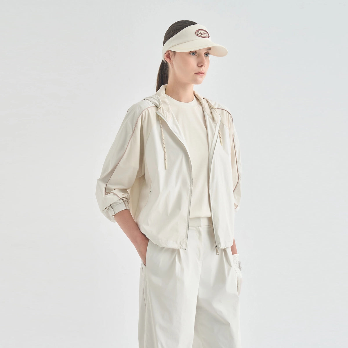 A model wearing a light beige hooded jacket, matching beige pants, a cream t-shirt, and a beige visor with a brown logo patch, standing against a solid light gray background.
