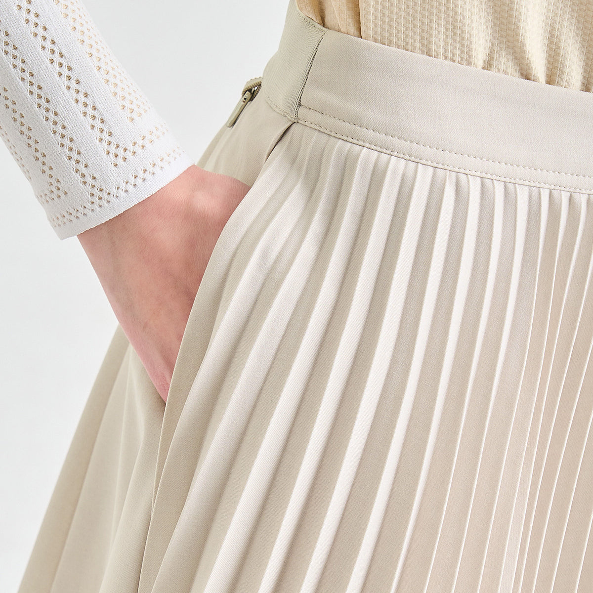 Close-up of a pleated high-waisted skirt in a light beige or cream color, featuring a zippered side pocket and a matching waistband, worn with a white textured long-sleeve top.