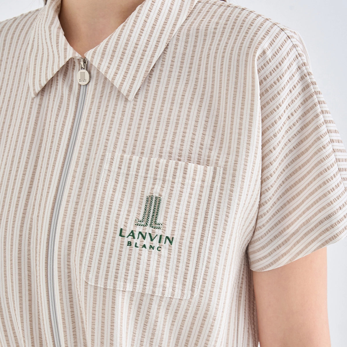 Close-up of a person wearing a short-sleeve collared zip-front shirt with vertical beige and white stripes and a green embroidered Lanvin Blanc logo on the chest pocket.