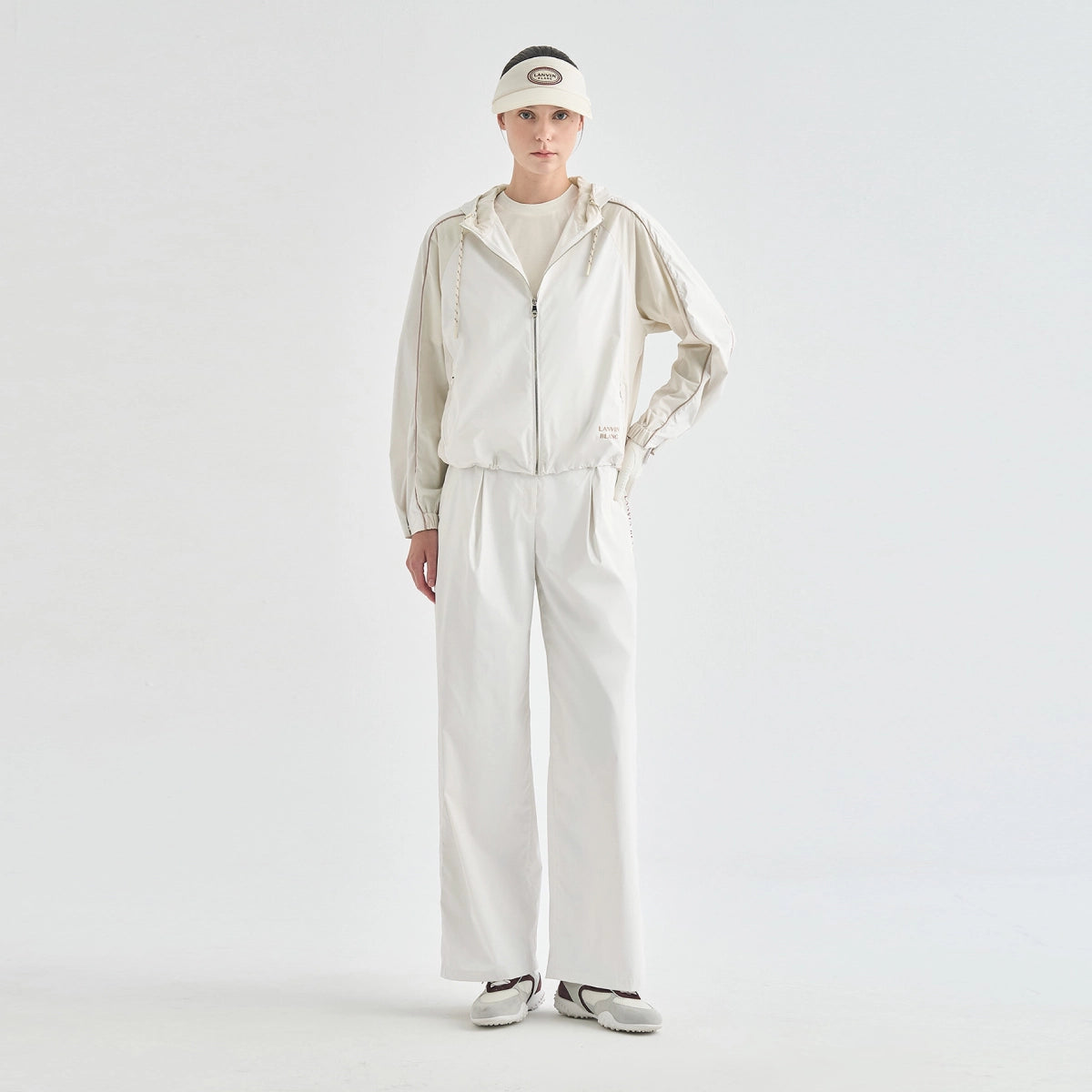 A model stands against a plain light gray studio background wearing a matching off-white tracksuit with a zip-up jacket, wide-leg pants, a visor, and sneakers.