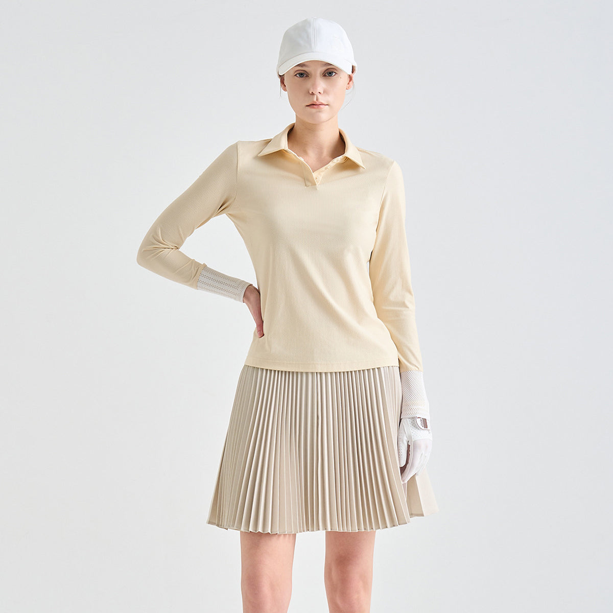 A woman modeling a beige long-sleeve collared golf shirt paired with a pleated beige golf skirt, white cap, and white glove against a light gray studio background.