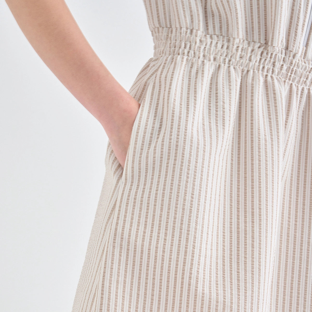 Close-up of a person wearing a cream-colored dress with thin vertical brown stripes and an elasticized waist, with a hand in a side pocket, against a plain white background.