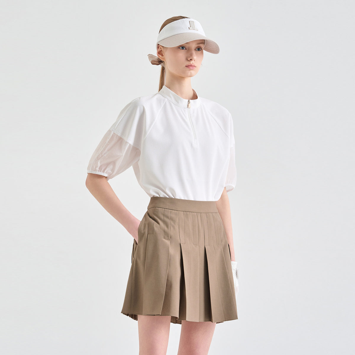 A model wearing a white short-sleeve golf polo shirt with sheer puff sleeves, a beige pleated tennis skirt, and a white visor with a logo, set against a plain light gray studio background.