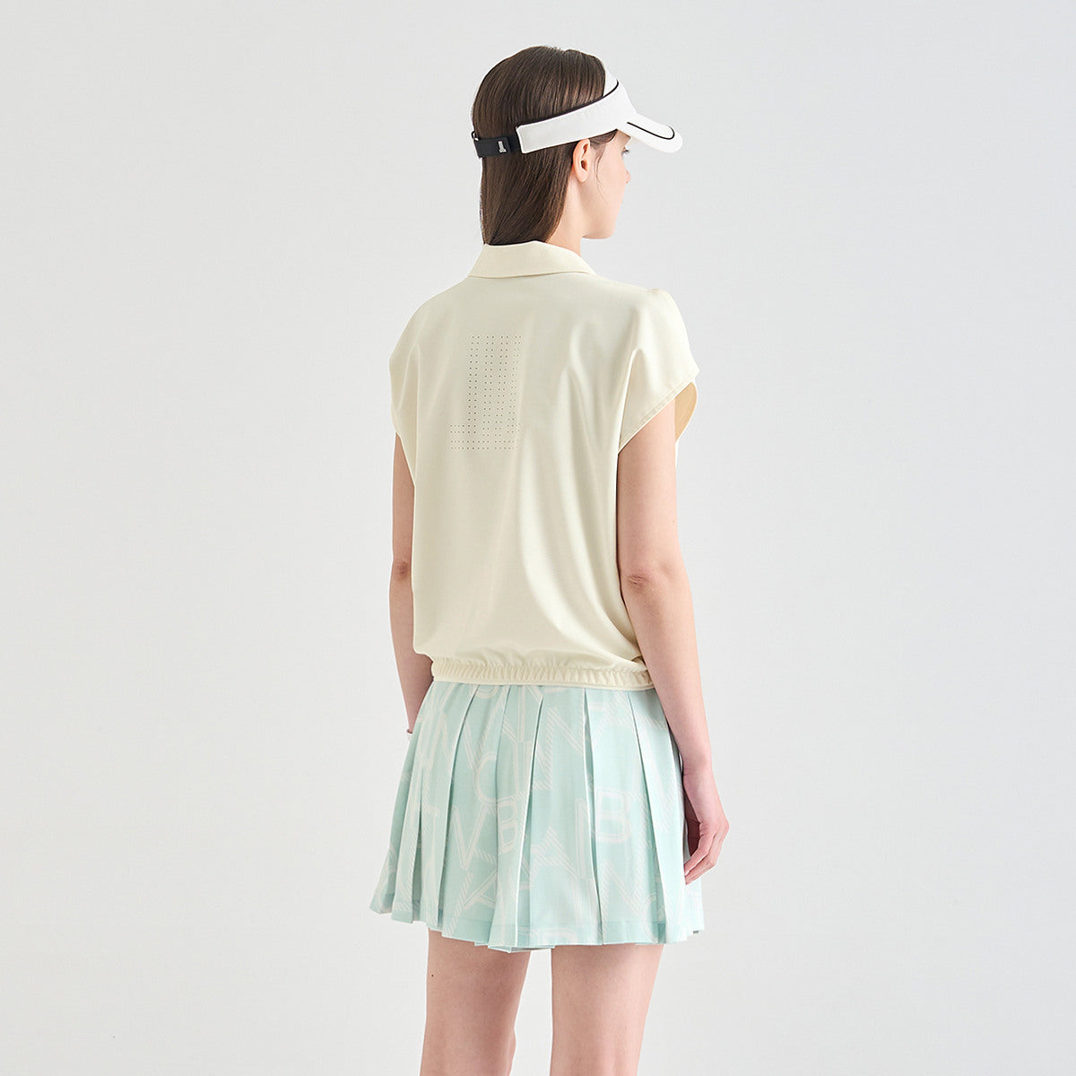 A woman shown from the back wearing a light cream sleeveless collared top, a mint green pleated tennis skirt, and a white visor, set against a plain light gray studio background.
