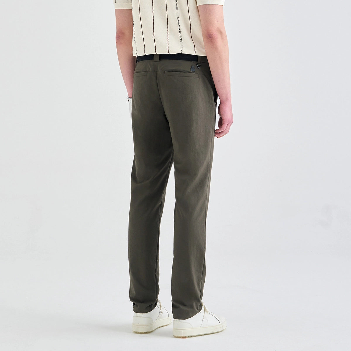 A person shown from the back wearing slim-fit olive green trousers, a cream short-sleeve shirt with vertical black stripes, and white sneakers against a plain light gray studio background.