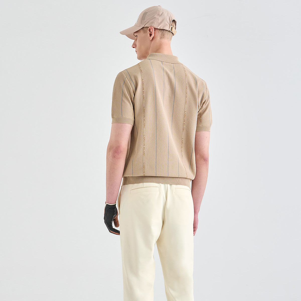 A man shown from the back wearing a beige short-sleeve knit polo shirt with vertical embroidered text, cream trousers, a beige cap, and a black glove, against a light gray studio background.