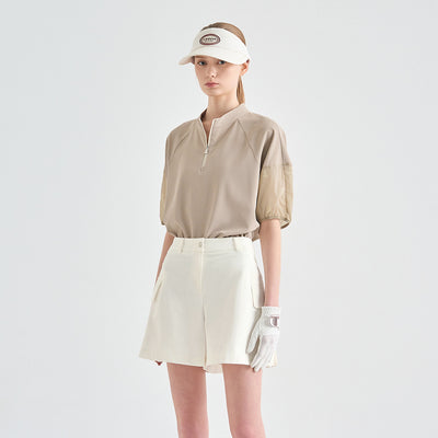 A model wearing a beige short-sleeve golf polo with textured puff sleeves, paired with cream shorts, a white visor, and a white glove, set against a plain light gray studio background.