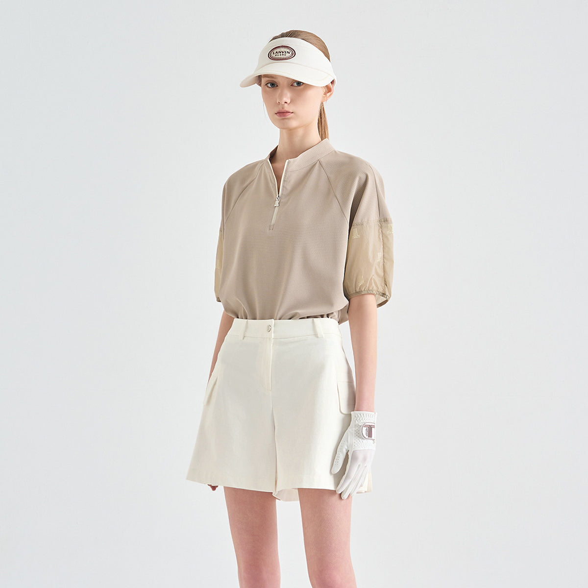 A model wearing a beige short-sleeve golf polo with textured puff sleeves, paired with cream shorts, a white visor, and a white glove, set against a plain light gray studio background.