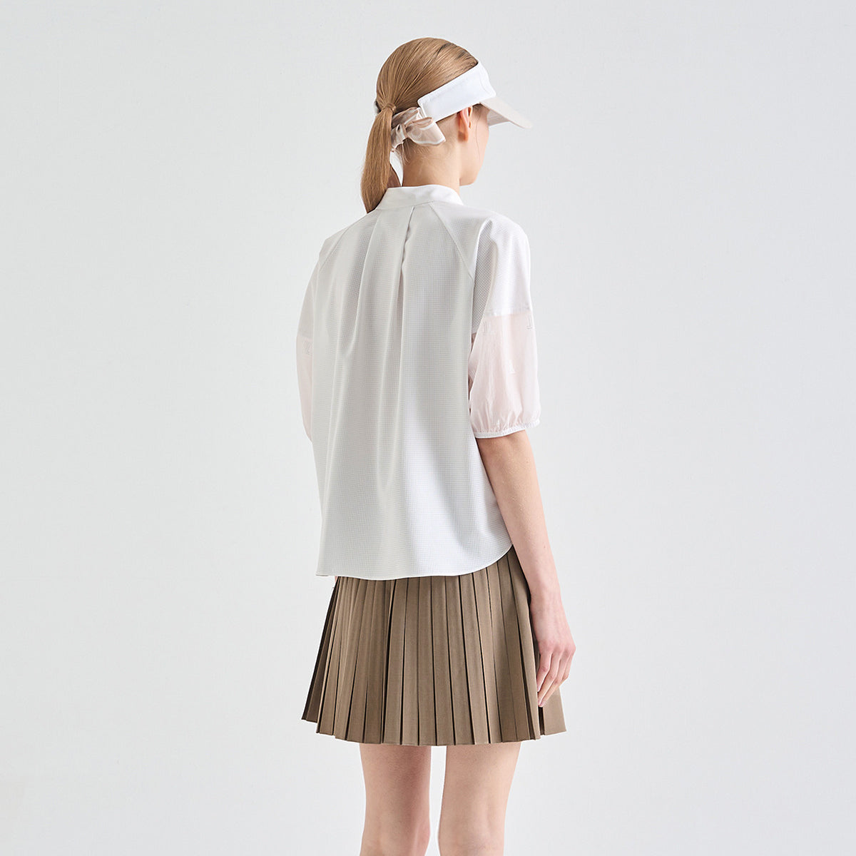 A woman shown from the back wearing a white short-sleeve blouse with sheer puff sleeves and a pleated taupe skirt, against a plain light gray studio background.