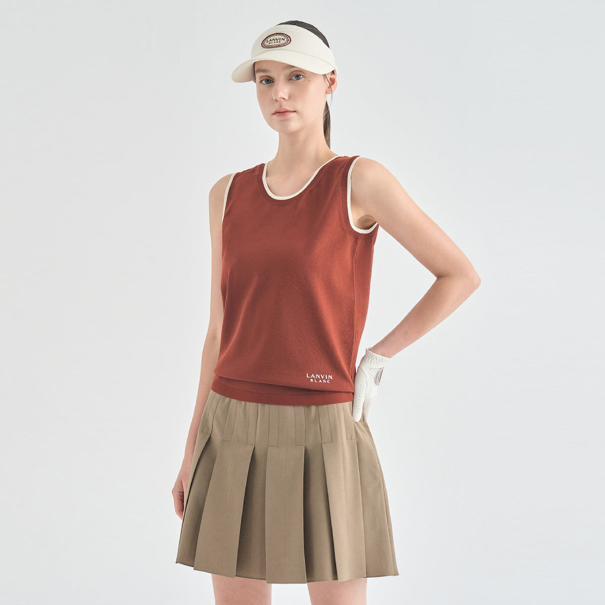 A woman modeling a sleeveless rust-orange athletic top with white trim, a beige pleated tennis skirt, a beige visor, and a white glove, against a light gray studio background.