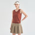 A woman modeling a sleeveless rust-orange athletic top with white trim, a beige pleated tennis skirt, a beige visor, and a white glove, against a light gray studio background.