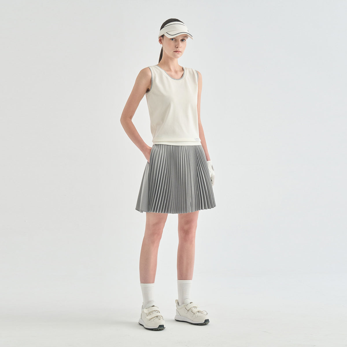 A model stands in a studio wearing a sleeveless cream top, a pleated gray tennis skirt, white socks, and white sneakers against a light gray background.